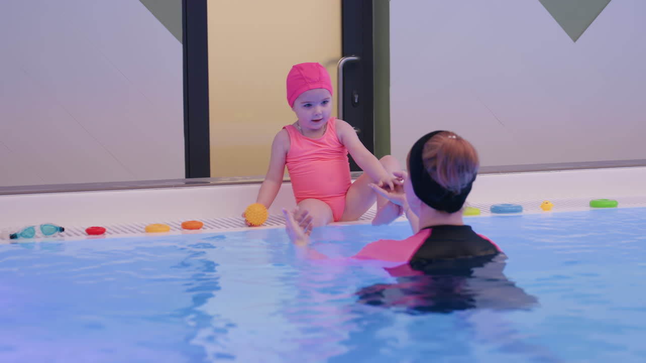 Infant taps small ball on rough pool pavement, exploring texture and sound, sunlight glints on wet tiles, splash nearby, curious hands test bounce, playful learning and coordination near clear water