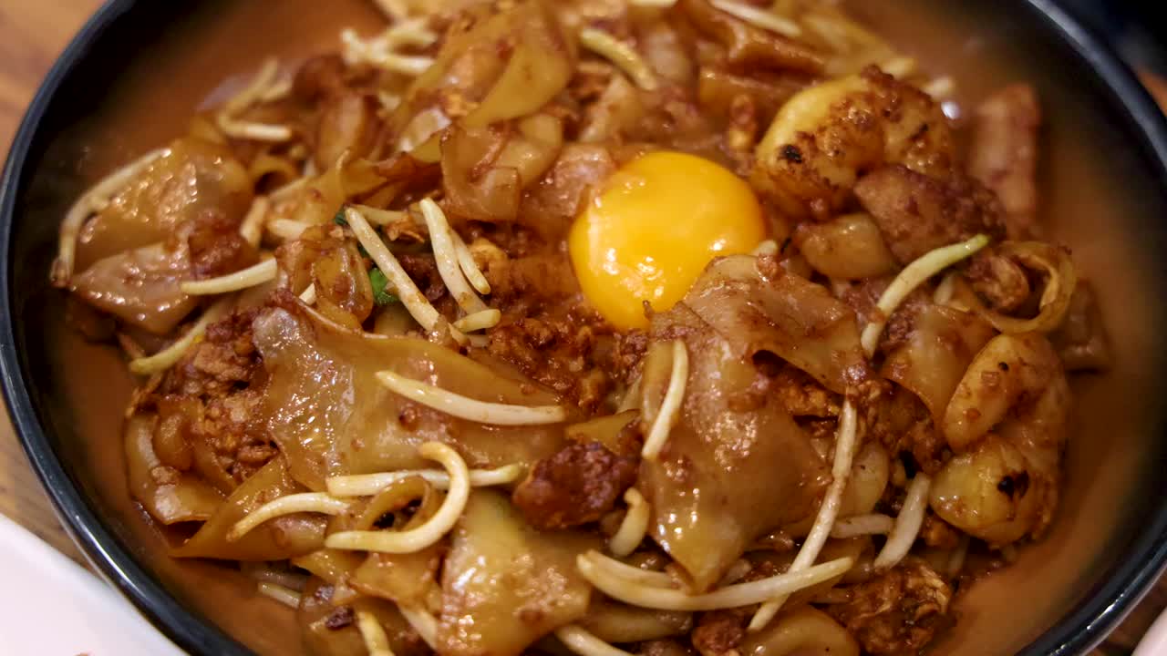 A bowl of stir-fried noodles with meat, vegetables, and a raw egg yolk is shown in close-up under warm lighting, with minimal camera movement