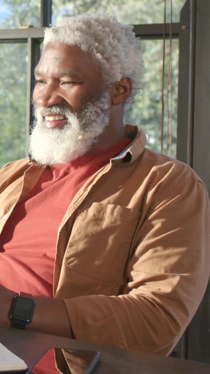 Vertical video: Leaning mature man wearing red shirt reading notebook by window, laughing