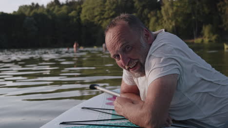 a happy elderly man leaning on a paddleboard smiles at the camera