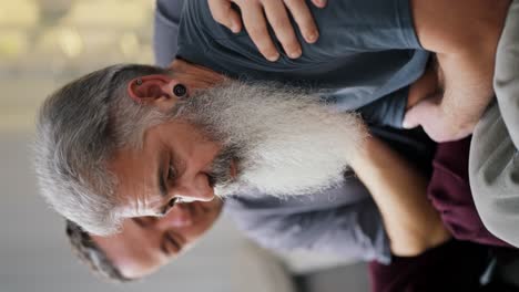 Vertical-video-of-a-sad-elderly-man-with-gray-hair-and-a-lush-gray-beard-in-a-gray-T-shirt-is-sad-and-looks-thoughtfully-while-sitting-on-the-sofa-with-his-boyfriend-a-middle-aged-brunette-with-stubble-in-a-modern-apartment