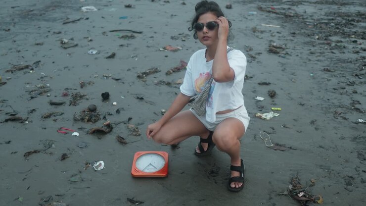 Giovane donna preoccupata accovacciata accanto a un orologio da parete arancione sommerso su una spiaggia di sabbia nera piena di spazzatura.