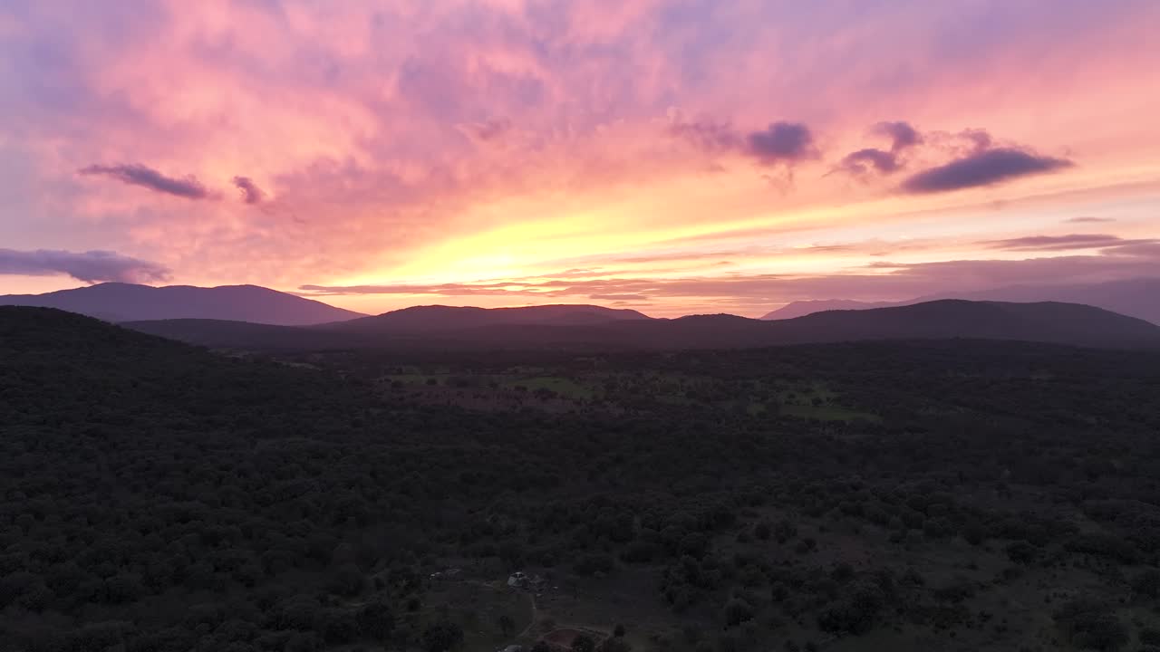 Drone ascent in a valley at dusk, with distant mountains and a sky full of colors—orange, yellow, blue, red, and violet. Clouds reflect rays, creating light lines as the day transitions to night.