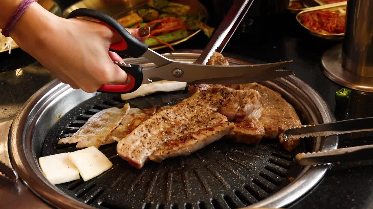 Hand cuts grilled pork belly with kitchen scissors on tabletop Korean barbecue grill, warm lighting