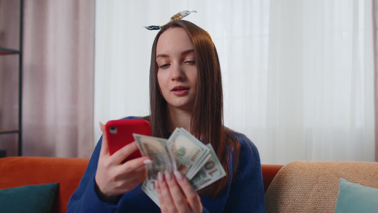 улыбающаяся счастливая молодая женщина с наличными деньгами и мобильным телефоном рассчитывает домашние счета дома