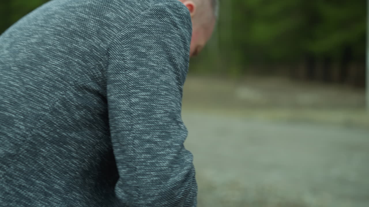 회색 재을 입은 남자가 철도 트랙 에 앉으려고 할 때 권총을 들고 있는 가까운 모습, 그는 초록색 숲의 보케 뷰와 함께 집중된 표정으로 권총을 검사하고 있습니다.