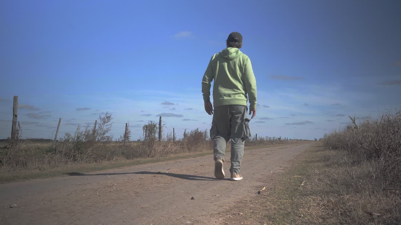 Wide view of a man walking away from the camera over a dirt road in a rural setting