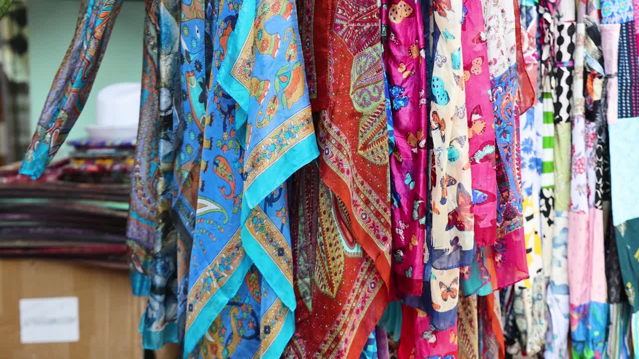 Vibrant silk scarves gently moving on rack, natural daylight, handheld camera, Singapore market setting