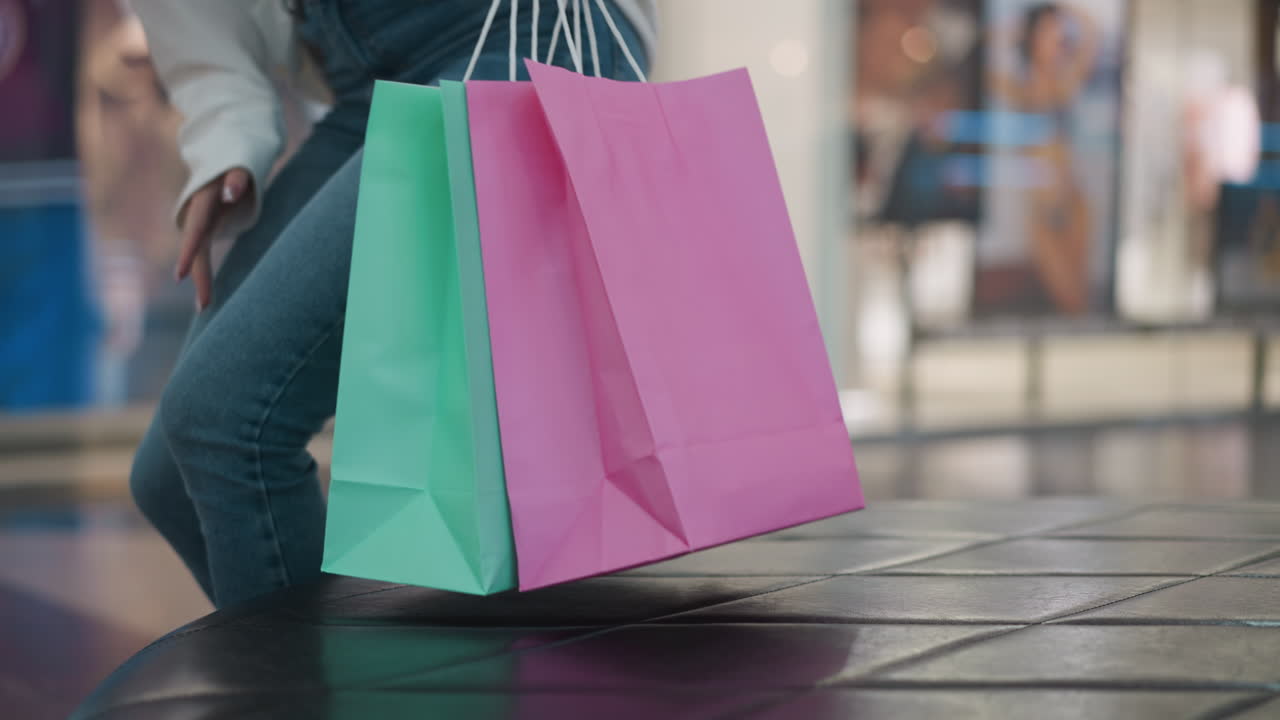 mujer se sienta con bolsas de compras en el centro comercial, luego se levanta y lleva las bolsas a mano, alejándose del asiento de cuero mientras el ambiente vibrante del centro comercial es visible en el fondo