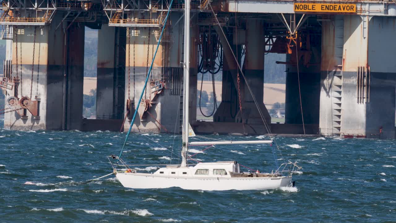 White sailboat moves past large offshore oil platform on choppy sea, daylight, telephoto shot