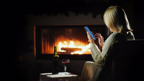 nice evening by the fireplace a woman uses a smartphone near a glass of wine