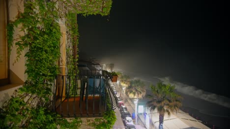 Static shot of a balcony covered with green plants, with street lights below and ocean waves crashing in the background at night, Letojanni, Taormina, Sicily, Italy (Italia)