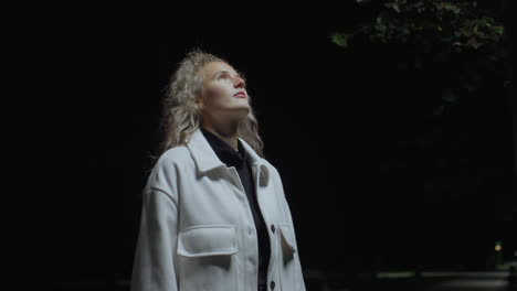 white woman wearing white coat stands under street light at night, gazing upward with calm expression, dim surroundings and gentle shadows create quiet, thoughtful atmosphere in night time park