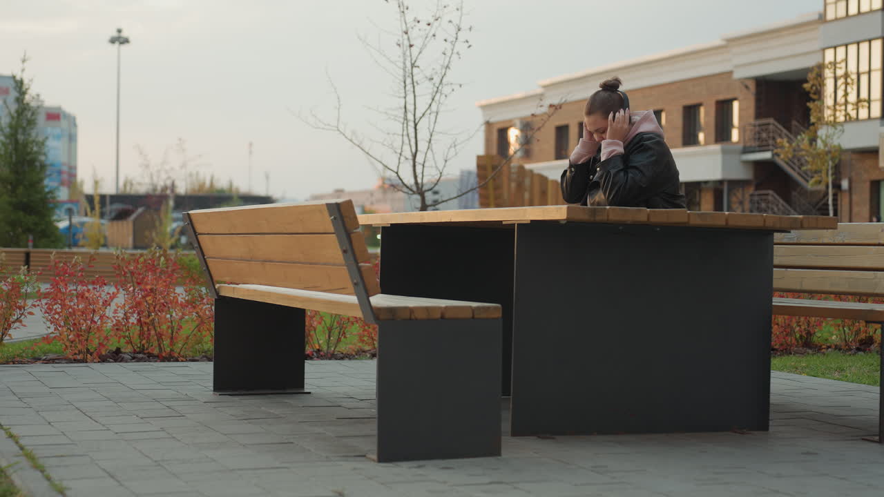 Young lady wearing leather jacket seated at wooden table outdoors putting on headphone with buildings in background and red shrubs swaying gently in wind under overcast sky
