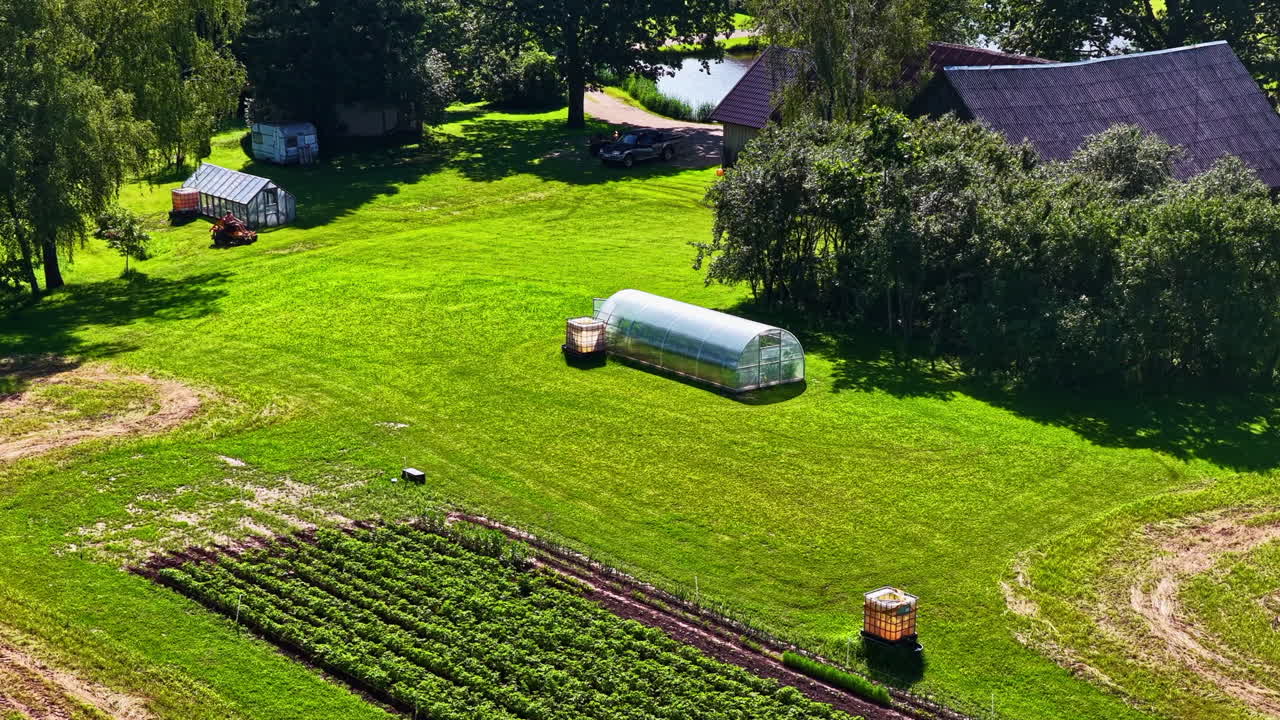 Aerial timelapse of a rural farm as a mower cuts grass across a bright green field. Active countryside scene with greenhouses and farmland