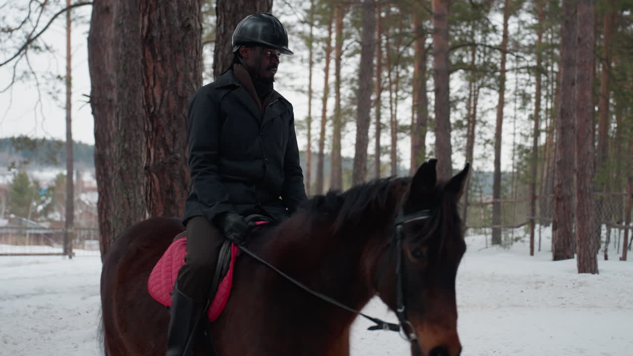 눈 덮인 소나무 숲을 말을 타고 달리는 흑인 남성, 좁은 길을 따라 일정한 속도로 걸으며 저 멀리 마을의 모습이 보입니다. 헬멧을 쓰고 겨울 장비를 착용한 기수와 갈색 말이 일정한 리듬으로 움직입니다.