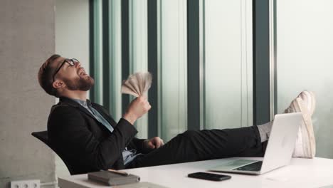 Un-Hombre-Exitoso-Sentado-En-Una-Mesa-Con-Una-Computadora-Portátil-Agitando-Billetes-De-Dinero