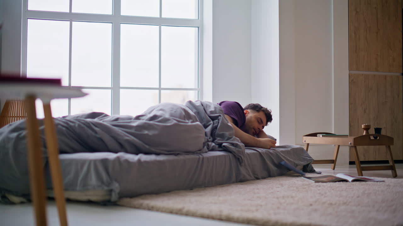 Søvnig mand vågner af alarmen og taber smartphonen tidligt om morgenen. Fyr vågner på madrassen.