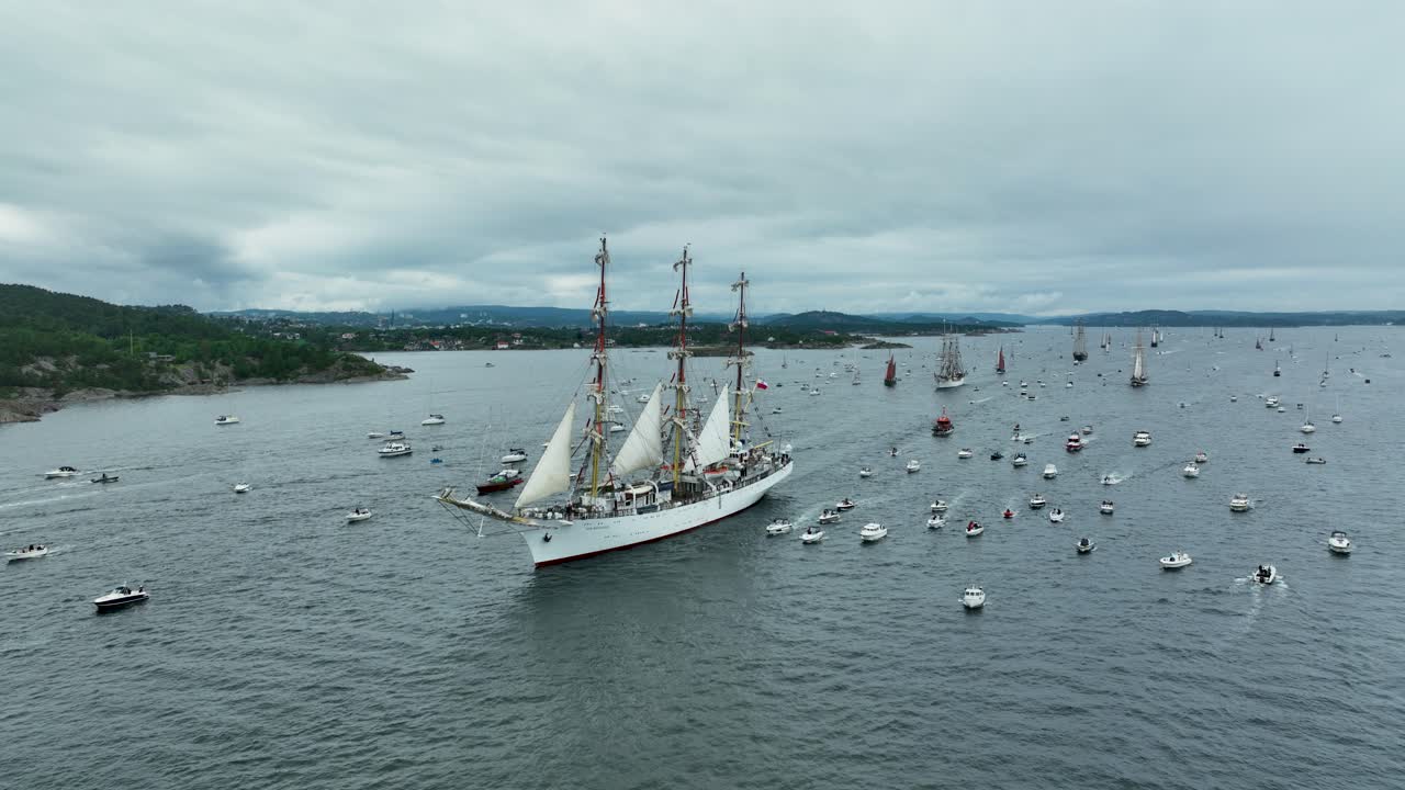A majestic tall ship sails gracefully among a multitude of smaller boats during a maritime festival on calm waters. The overcast sky adds a dramatic backdrop to the event