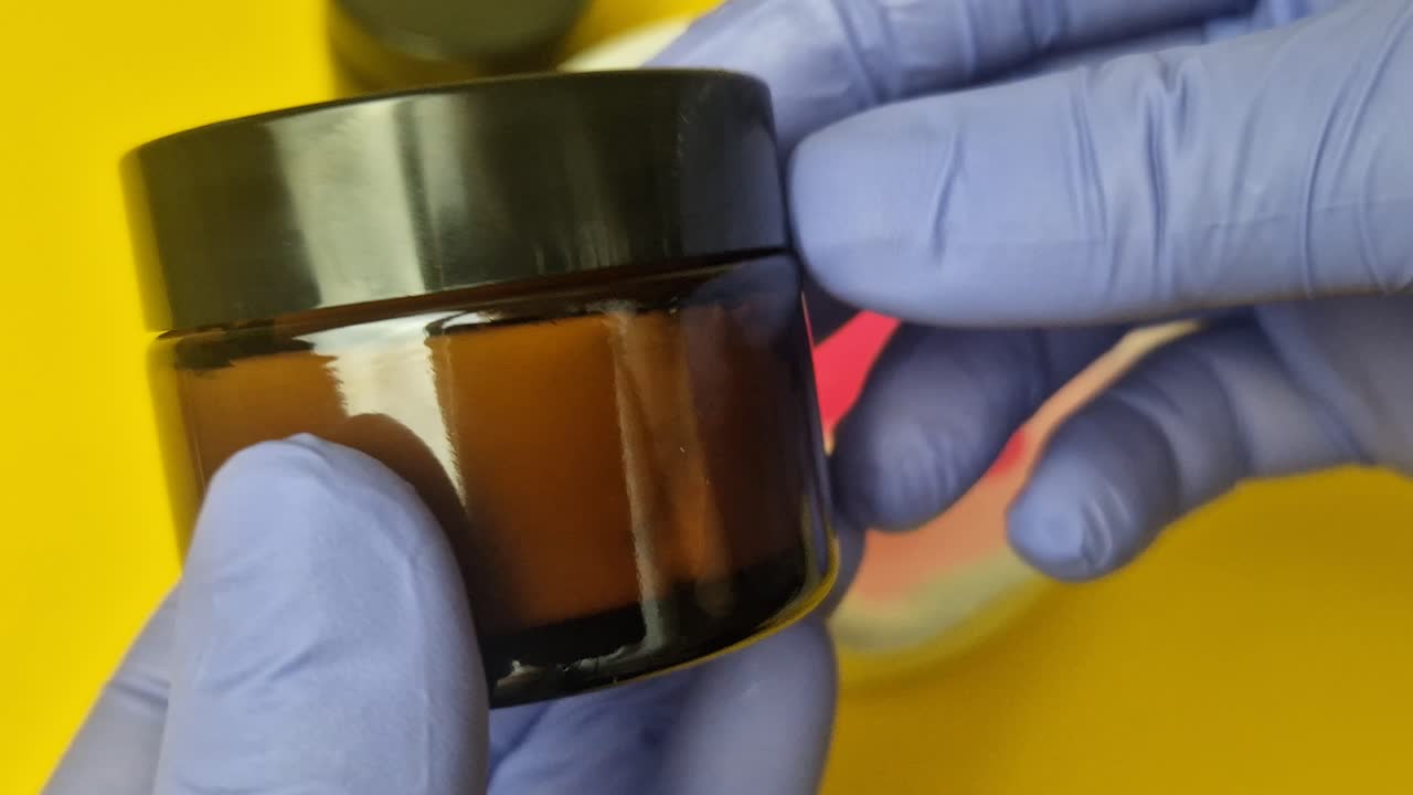 Close-up of gloved hand holding a black jar filled with freshly formulated cosmetic cream. Focus on the rich, yellow texture. Ideal for beauty, DIY, and pharmaceutical concepts