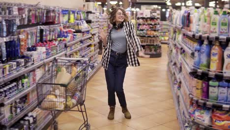 Full-length-footage-of-young-woman-in-jeans-and-plaid-shirt-dancing-standing-at-grocery-store-aisle.-Excited-with-headphones-on