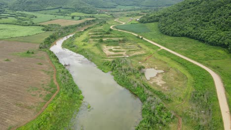 Farmlands-and-Mountains-in-Muak-Klek,-Thailand,-a-reverse-aerial-footage-revealing-this-farm-road-on-the-right-and-the-muddy-flowing-river-in-the-middle