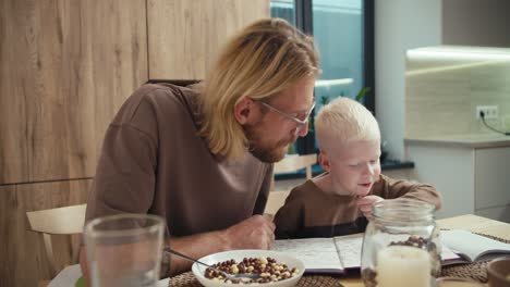 A-little-albino-boy-with-white-hair-reads-a-book-with-his-father,-a-blond-man-with-glasses-and-a-beard,-in-the-kitchen-after-breakfast-in-the-morning-in-a-modern-apartment