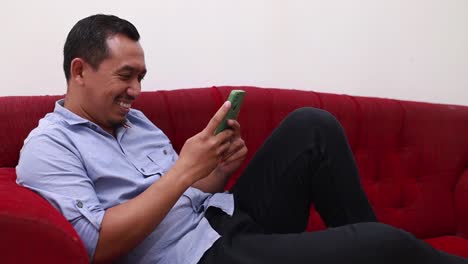 Glücklicher-Asiatischer-Mann,-Der-Sein-Handy-Benutzt,-Während-Er-Zu-Hause-Auf-Dem-Roten-Sofa-Sitzt