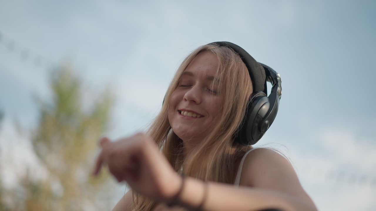 Giovane donna caucasica con le cuffie che balla all'aperto sotto delle luminarie, occhi chiusi e sorriso, gesti ritmici con le mani, atmosfera estiva rilassata, energia da ballerina di strada, aria da studentessa, frequentatrice di festival