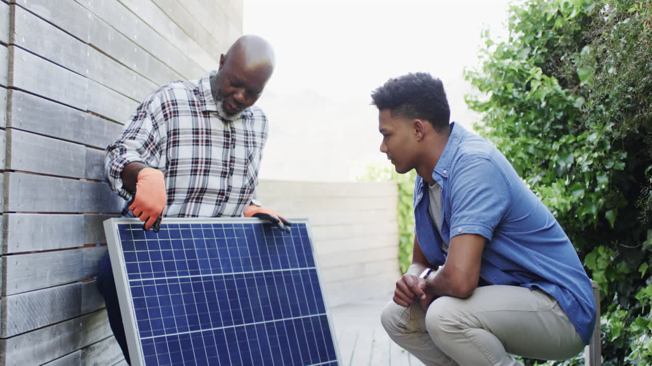 African american father showing solar panel to adult son in sunny garden, slow motion