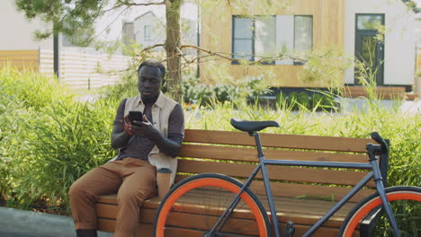 Hombre-Afroamericano-Usando-Un-Teléfono-Inteligente-En-Un-Banco-En-El-Parque