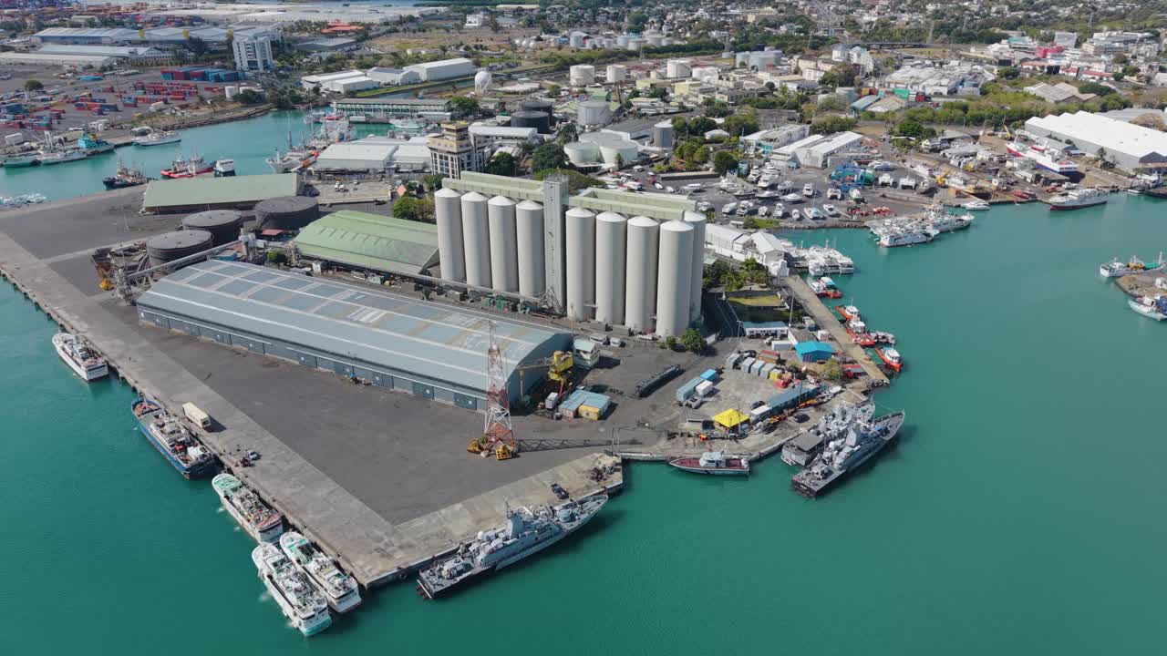 Slow orbiting drone shot of the Port Louis harbor in Mauritius, featuring large industrial grain silos, warehouses, and docked ships against the backdrop of the city and blue water