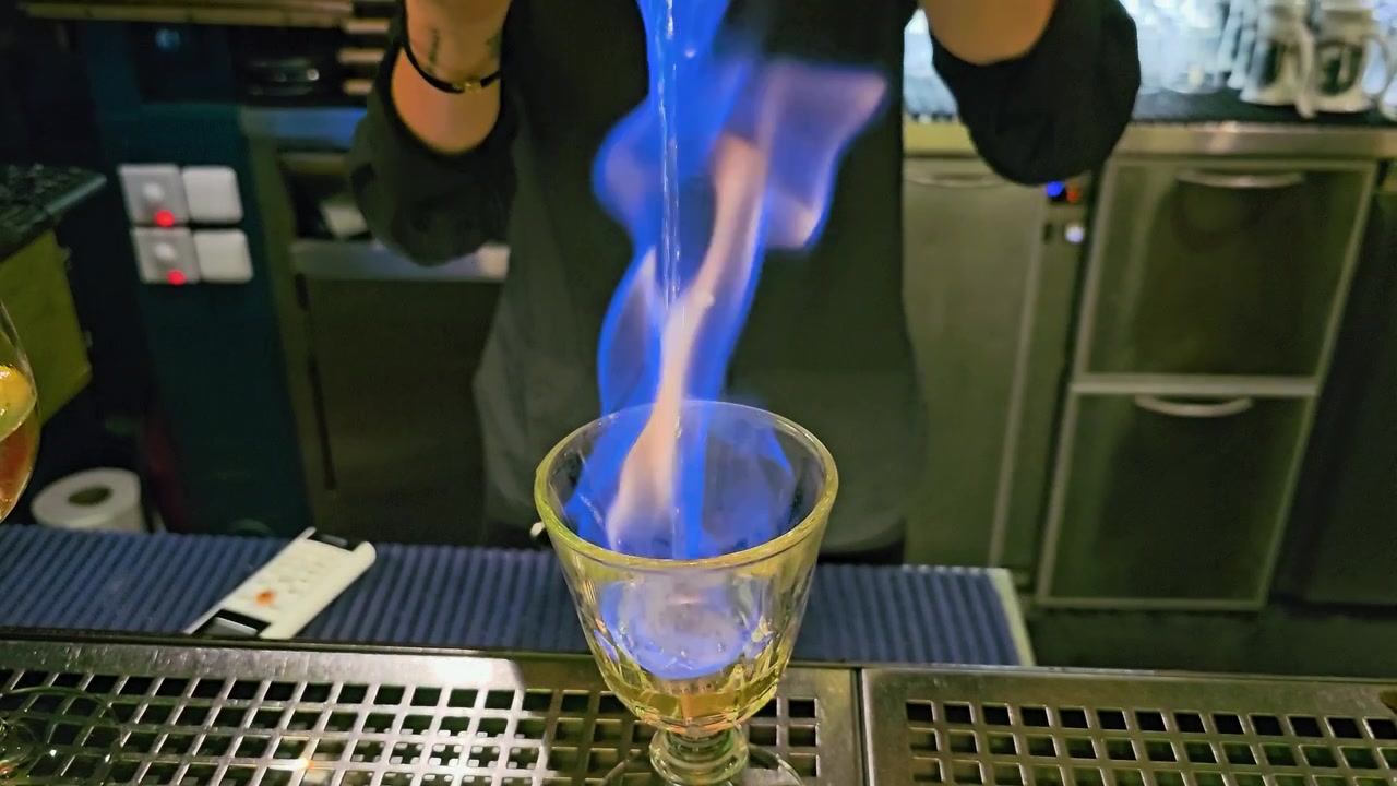 Primo piano di un barista che versa un cocktail fiammeggiante in un calice di vetro, con fiamme blu e arancioni, shaker in metallo, bancone del bar in acciaio inossidabile e un drammatico servizio da discoteca al chiuso di notte