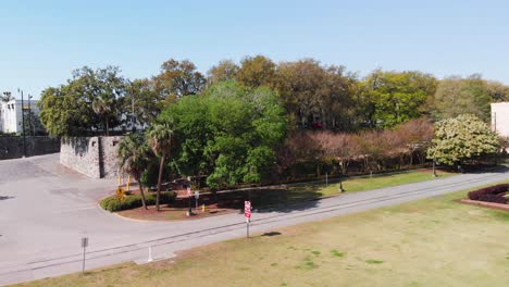 drone is rising up over green park area with lots of trees and street traffic visible in background