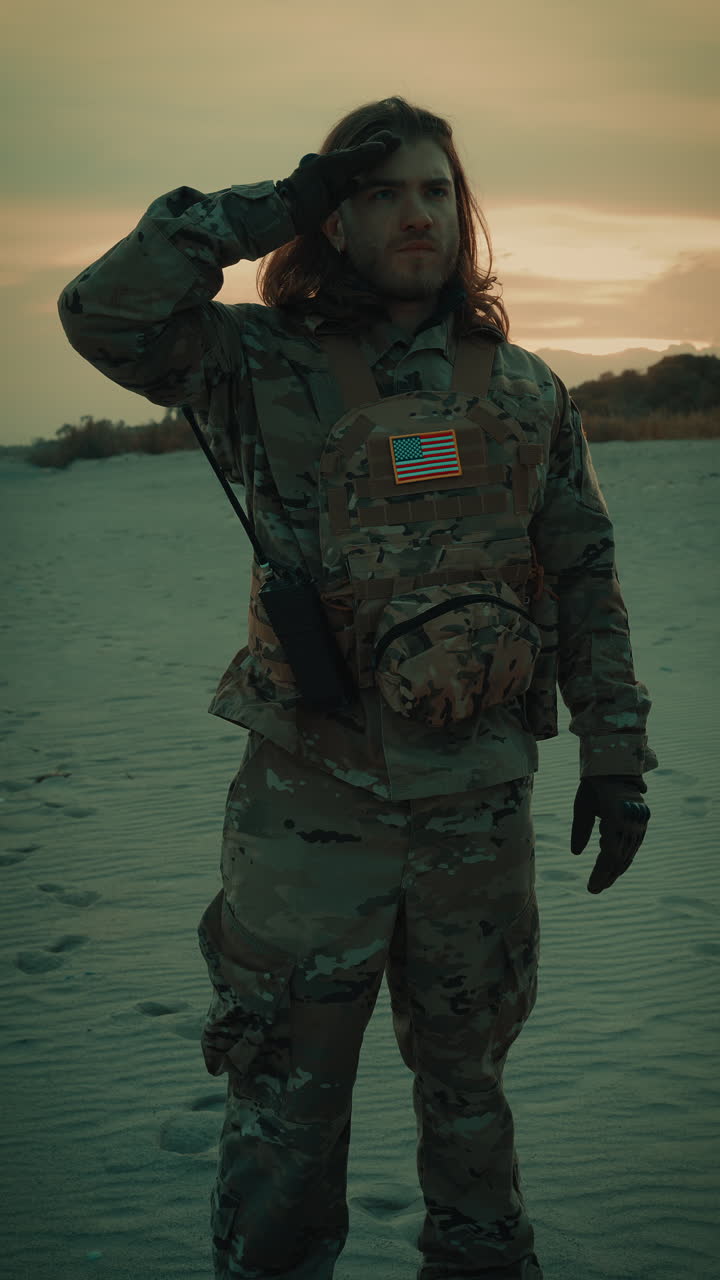Soldato americano con capelli lunghi che saluta di fronte alla bandiera sulla spiaggia al tramonto