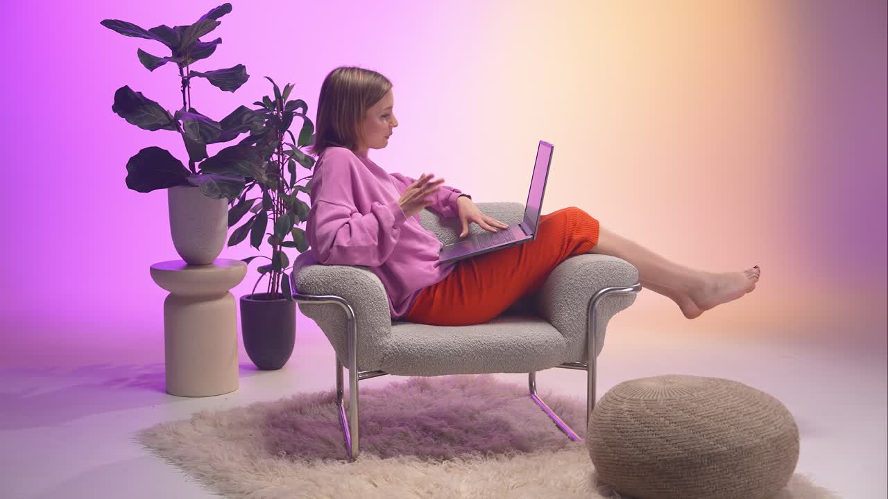 A young woman in a pink sweatshirt and orange pants works remotely on her laptop, barefoot in a stylish chair surrounded by pastel lighting, lush houseplants, and modern home decor