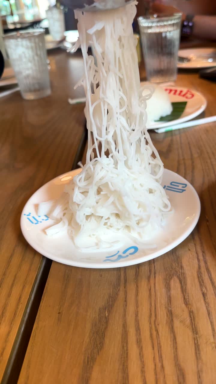 Una mano solleva i noodles di riso Kanom Jeen dal piatto in un luminoso ristorante di Bangkok, con luce naturale del giorno, primo piano.