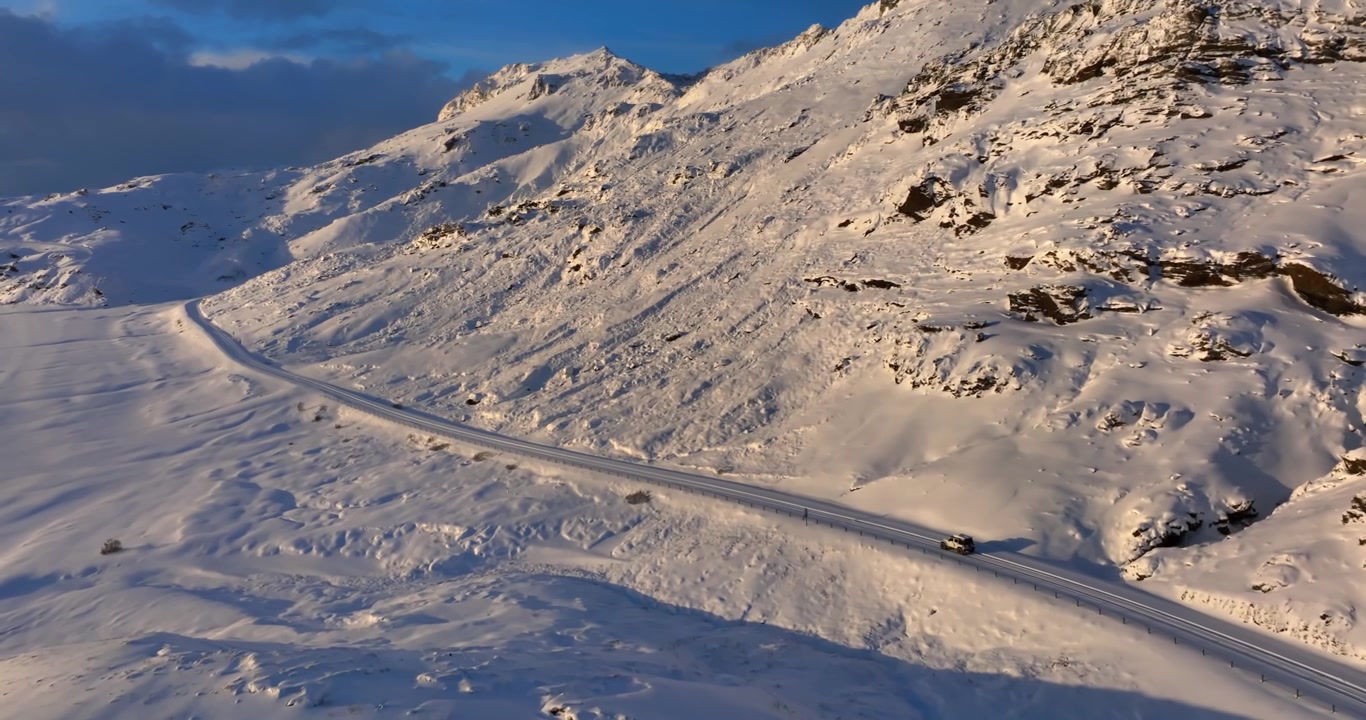 Aerial view of a lone vehicle driving along a snow-covered mountain road near Kleifarvatn, with sunlit volcanic slopes and vast Icelandic winter terrain.
