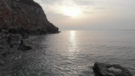 as sun rises on horizon of calm sea, low-tracking drone shot glides along rocky coastline