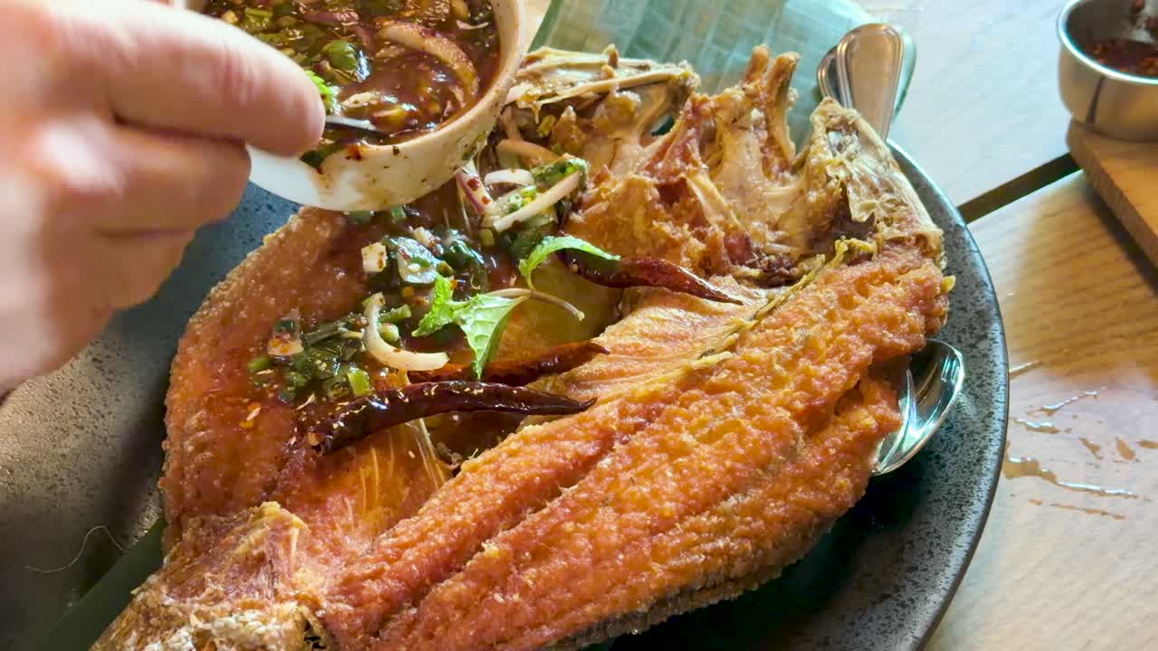 Hand pours chili herb sauce onto crispy fried sea bass in bright, natural restaurant lighting