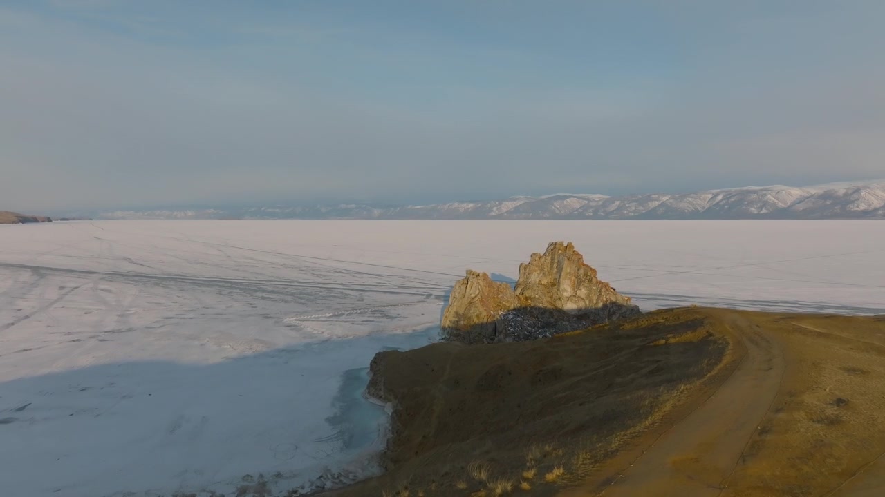 Droneoptagelse af Shamanka-klippen på Olkhon-øen, omgivet af den frosne Bajkalsø, Irkutsk oblast, Rusland, med sneklædte bjerge i horisonten.