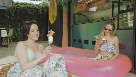 two young women enjoy ice cream while relaxing in an inflatable pool in front of a modern ice cream parlor with tropical decor.