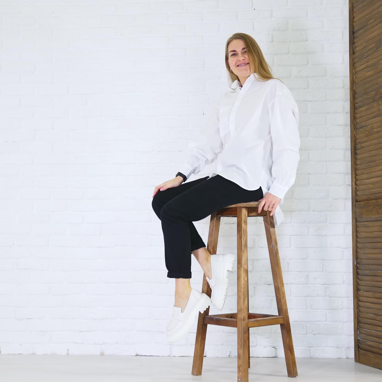 Smiling lady sitting on high stool and waving legs. Demonstrating new white shoes at the backdrop of white wall and wooden doors