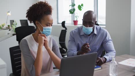 Diverse-businessman-and-businesswoman-in-face-masks-discussing,-using-laptop-in-office