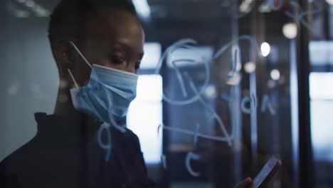 african american woman wearing face mask writing with marker pen on on glass board at modern office