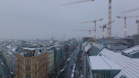 Group-of-tall-tower-cranes-working-on-construction-site-of-new-multistorey-building-in-urban-neighbourhood.-Fly-above-street-in-winter-city.-Berlin,-Germany