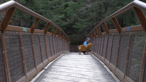 Young-woman-hiker-and-her-dog-stop-take-a-break-in-the-middle-of-a-metal-bridge-to-take-in-the-view-of-the-river-below