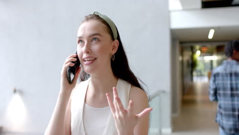 talking on smartphone, woman gesturing with hand in modern office hallway