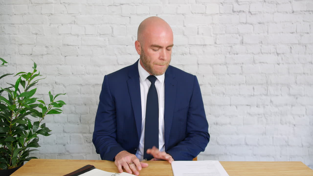 A bored businessman is fidgetting at his desk, The bald man in a suit is tapping his fingers on the desk as he drums a beat