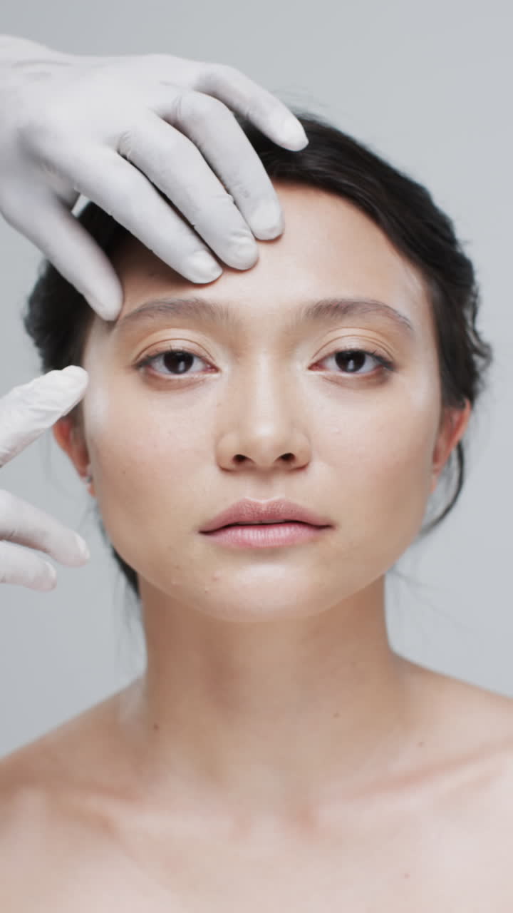 Vertical video of touching face of asian woman on grey background, slow motion